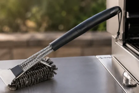Cuisinart-style grill cleaning brush with metal bristles and scraper resting beside an outdoor barbecue grill.