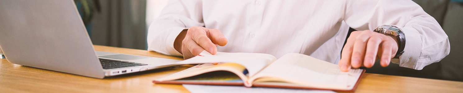 A person looking at a book in front of a laptop on a desk