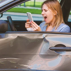 A woman looking at the phone inside a car with a dent on the side