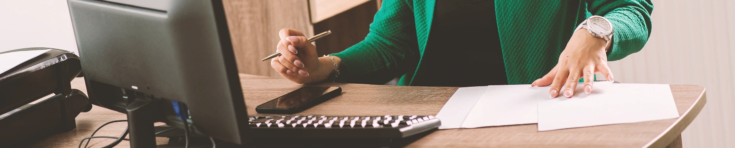 A person doing paperwork while in front of a computer and using a phone