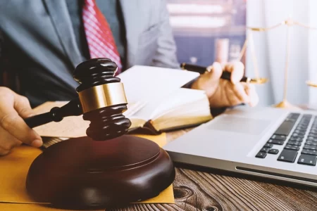 A lawyer at his table in front of his laptop while holding a book and a gavel